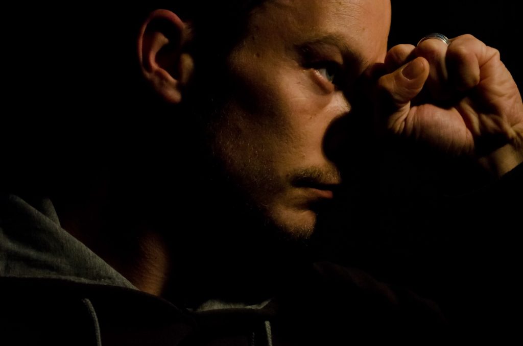 Somber closeup photo of a man in the shadows resting his head on his hand. He's of indeterminate race, we see him from the side. 