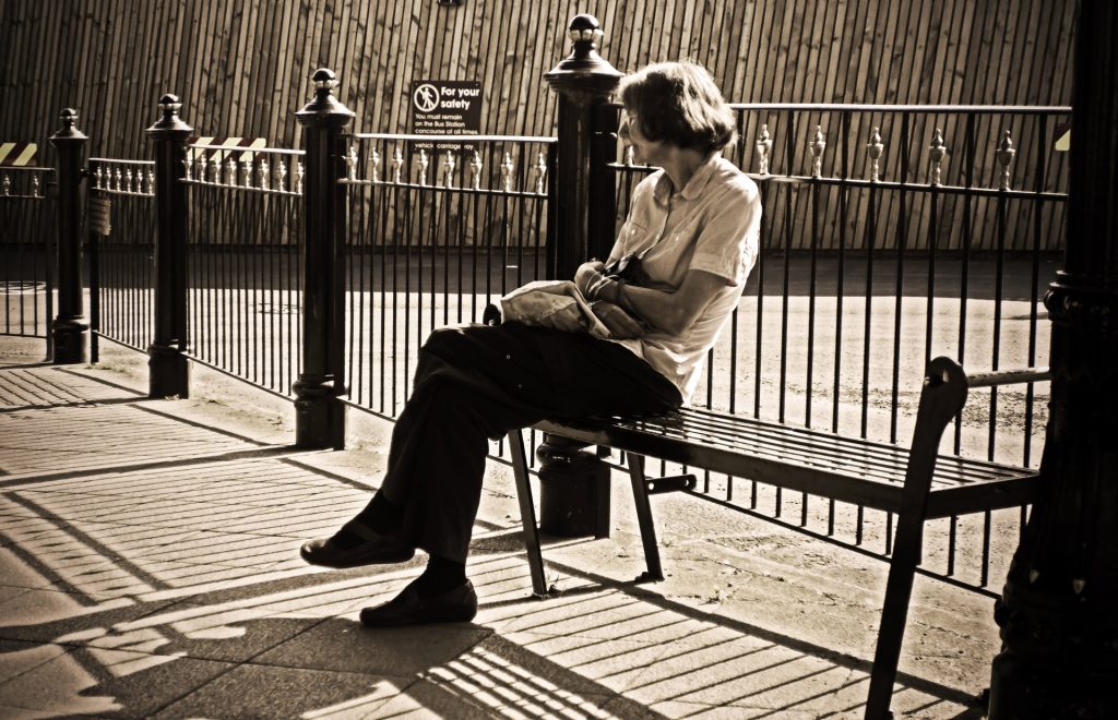 Sepia toned middle aged short haired woman with dark hair and a bag and pants and shoes waiting alone on a bench near a fence. 