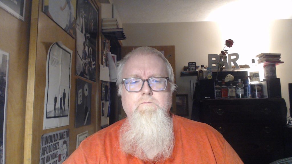 Older middle aged white man with a balding head, a white beard, reading glasses, and a bright orange tee shirt standing in a bedroom with a dresser and a rose and liquor bottles and a wall full of posters.