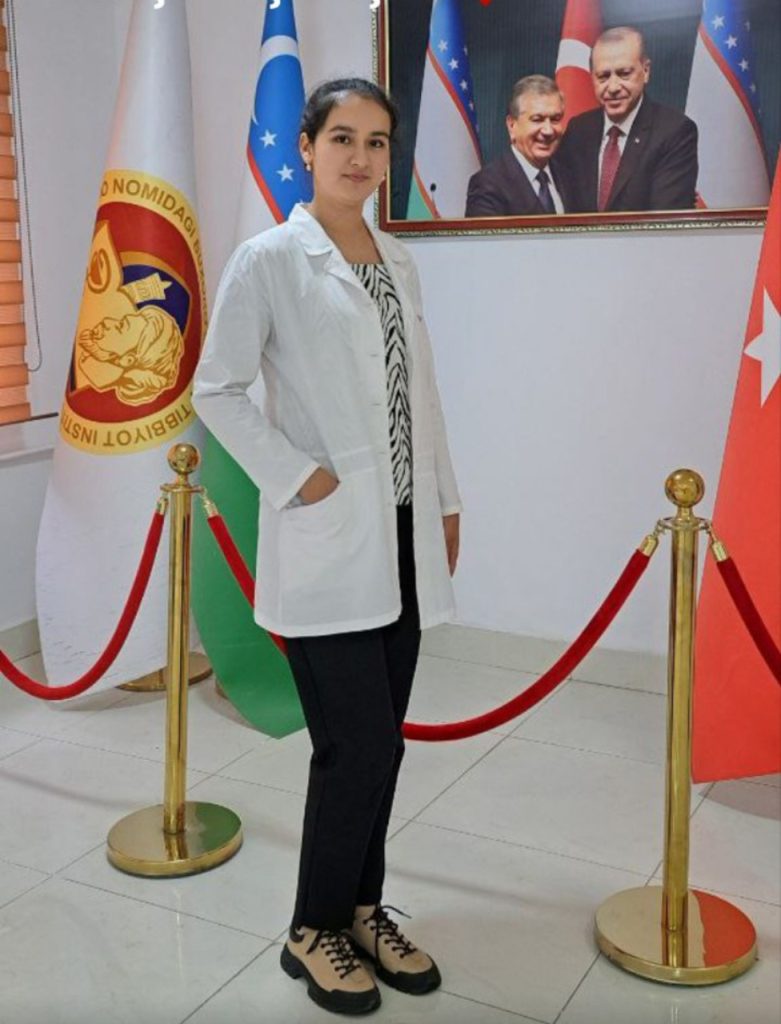 Central Asian young woman in a white coat, small earrings, a black and white top, and black dress pants stands on a stage near a photo of leaders of her country and flags. 