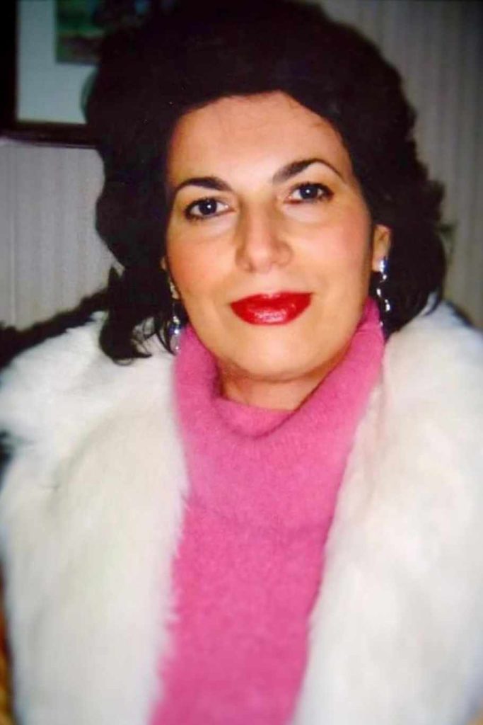 European woman with short dark curly hair, brown eyes, earrings, lipstick, a white fluffy coat and pink scarf. 