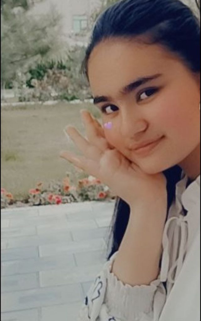 Central Asian teen girl peeking out at the camera with her head in her hands. She's got a painted floral scene in the background. 