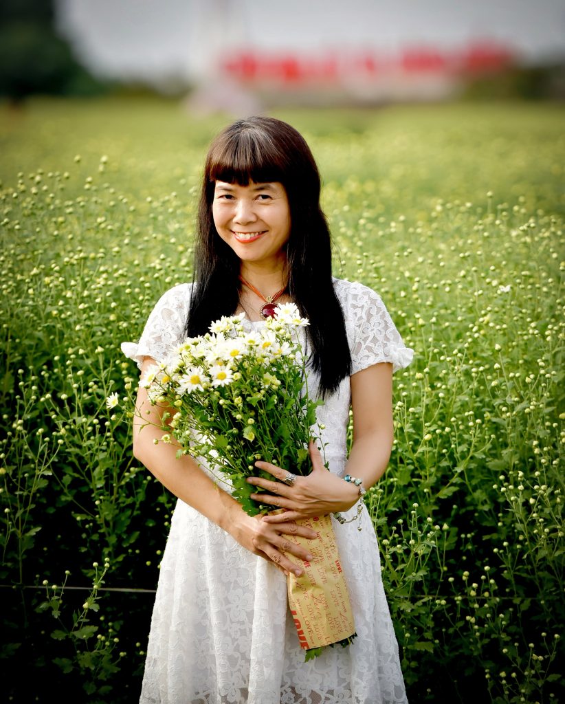 Young East Asian woman with long dark hair and brown eyes in a white lacy summer dress holds a bouquet of daisies in a field of them. 