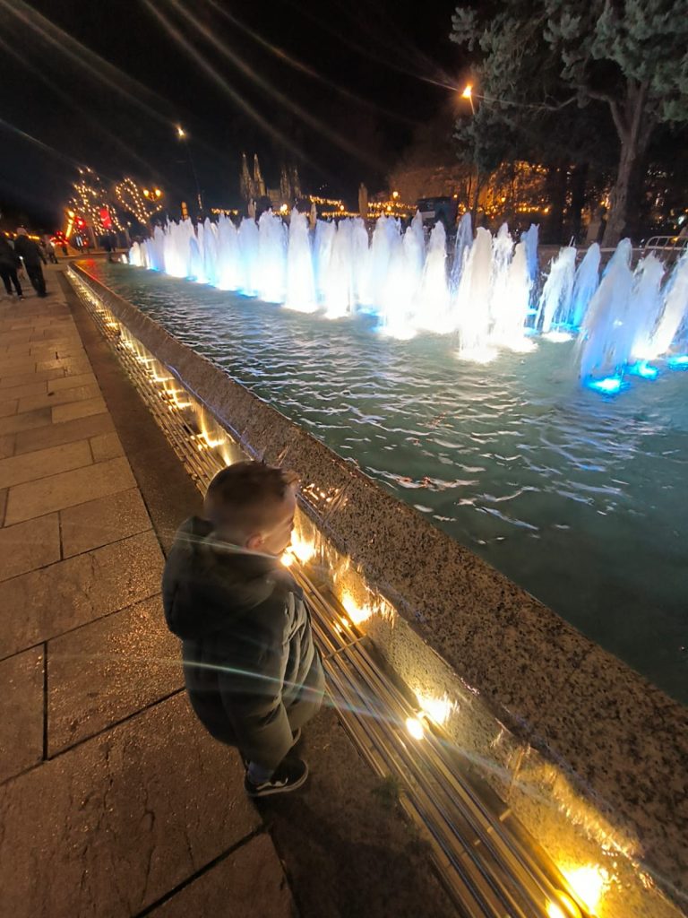 Young boy in a large puffy coat looks out at waterfall sculptures in a city fountain lit up at night. 