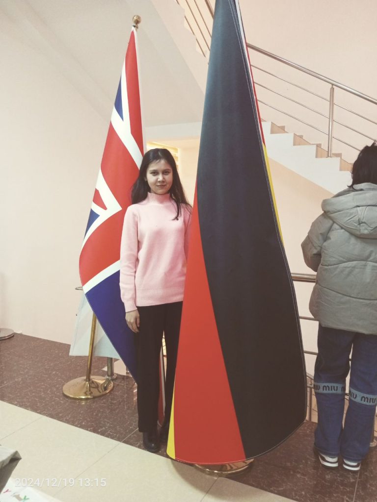 Central Asian teen girl with long dark hair in a pink sweater and black pants standing in front of flags and a stairwell. 