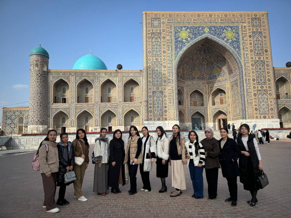 Group of Central Asian young women students in front of a decorated building with blue and yellow and tan designs, a few green domes, and arched entryways. 