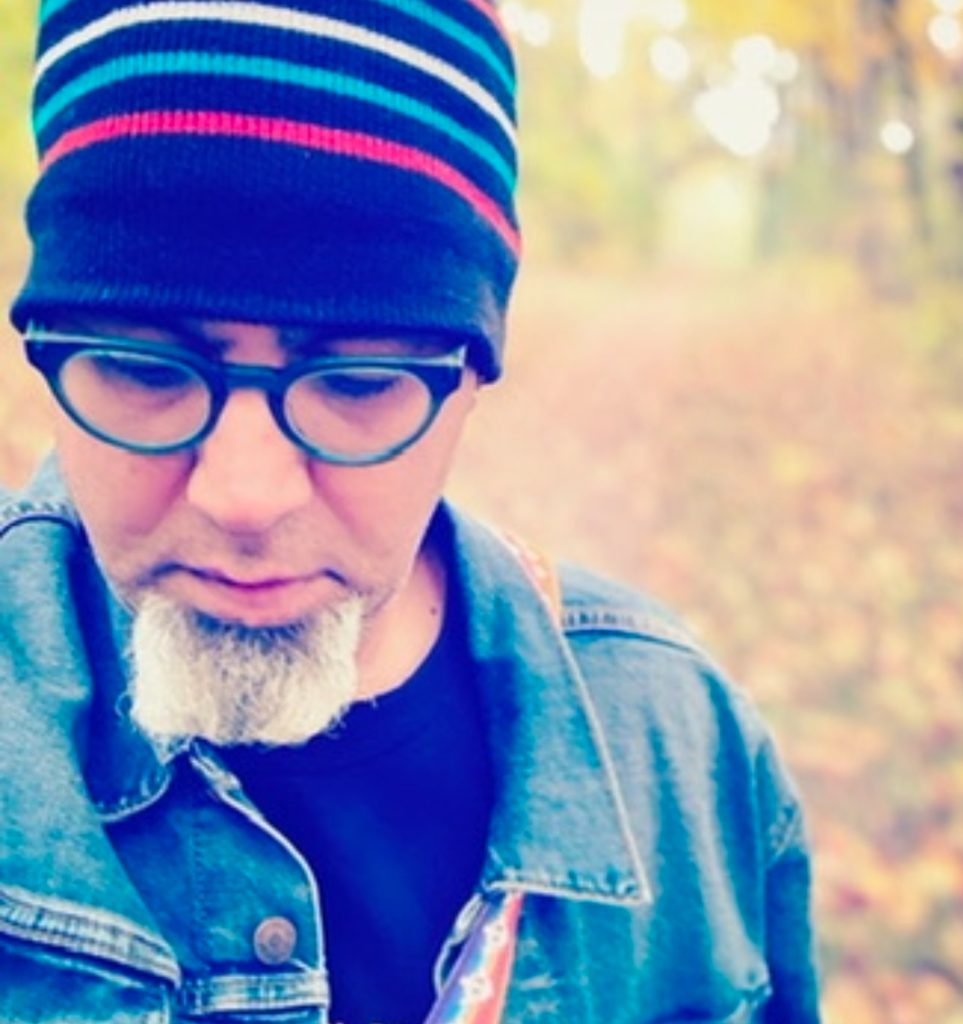 Young middle aged white man with reading glasses and a knit hat with stripes and a small beard and a jean jacket over a black top looking down. 