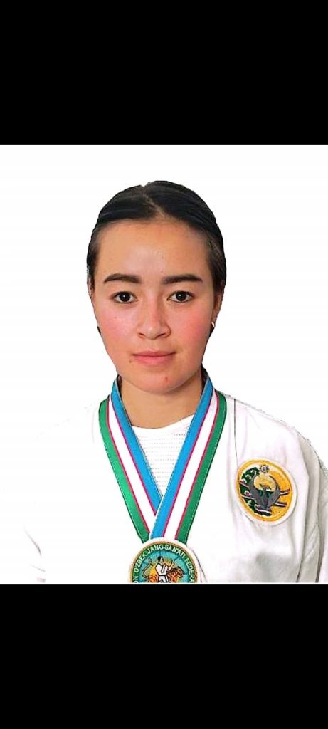 Central Asian girl with short dark hair, brown eyes, and a white long sleeved top with a patch on her breast and a medal around her neck. 