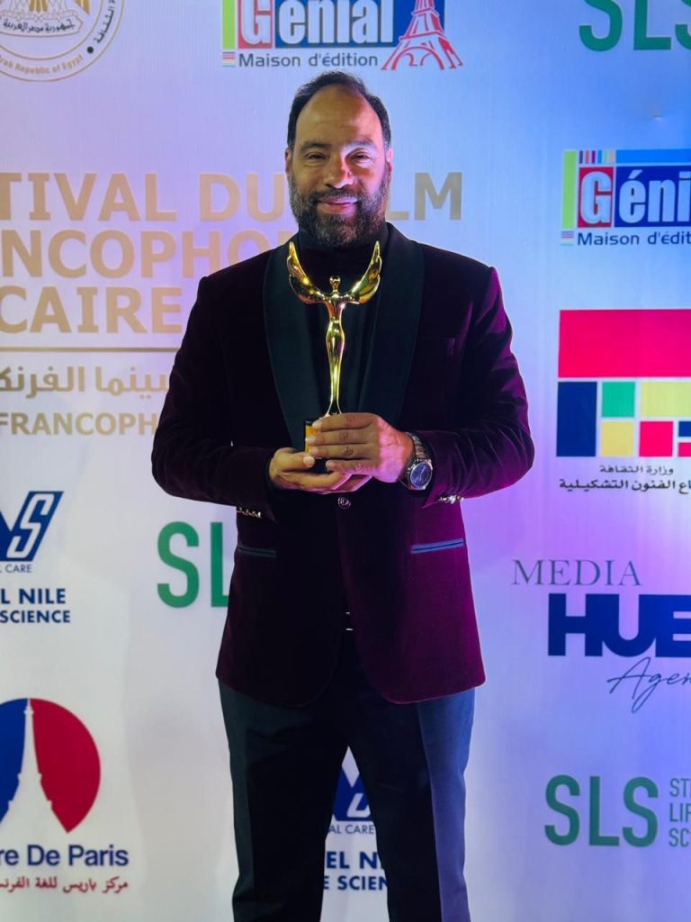 Egyptian man, older middle aged, short brown hair, mustache, and beard, in a dark suit holding a bronze award at a film festival. 