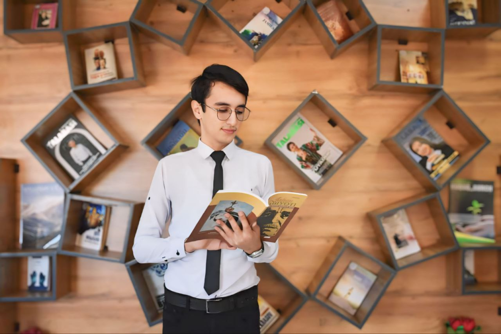 Young Central Asian teen boy in a white collared shirt and tie, with a belt and reading glasses and black pants. He holds a book open and has an artsy angled background of magazines positioned inside of blocks.