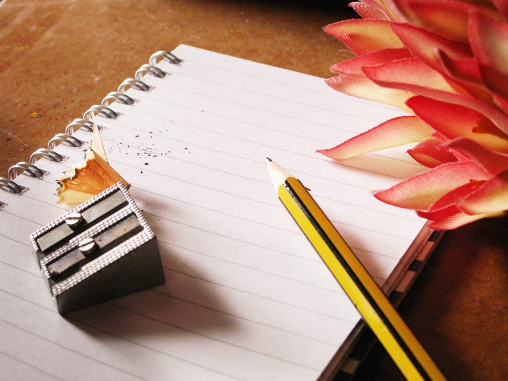 Yellow sharpened pencil, sharpener, pink flower and open notebook.