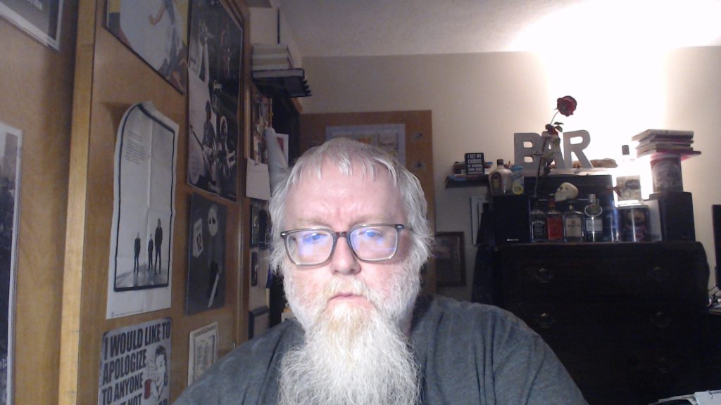 Older middle aged white man with reading glasses, a long beard, light blonde hair, and a gray shirt standing in a bedroom with a dresser and a rose and posters on the wall. 