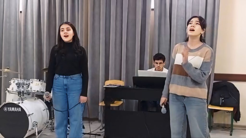 Two young Central Asian women in sweaters and jeans sing before a small group. They have microphones in their hands and a drum set and young man are behind them. 