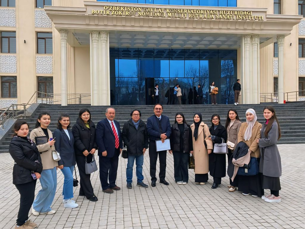 Group of students in front of a large building with glass windows and columns. Students are dressed up in coats, a few in headscarves, and the male teachers are in suits. 