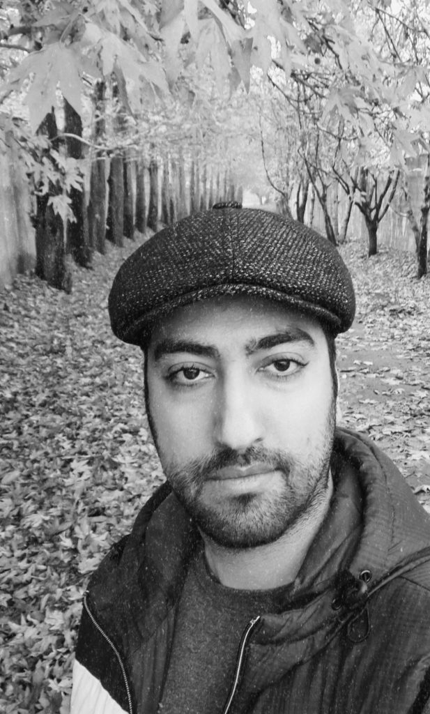 Black and white photo of a young Middle Eastern man with a beret, brown eyes, short hair, a jacket over a dark tee shirt. He's in a grove of trees with fall leaves. 