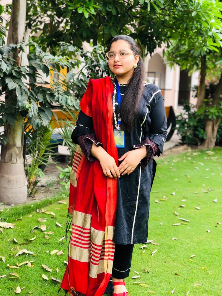 Young South Asian woman standing on a green lawn under leafy tree branches. She's in a black dress with white edges and a red scarf and a school ID around her neck, and has reading glasses and small earrings. 