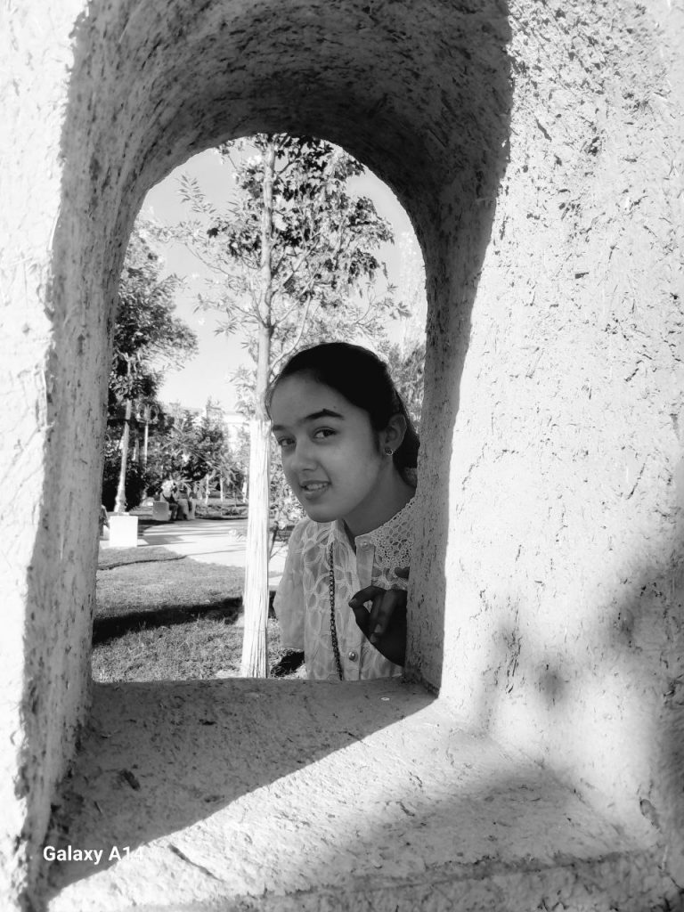 Black and white photo of a teen Central Asian girl with long dark hair and a white frilly blouse peering through an arched opening in a concrete wall. A park with trees is behind her. 