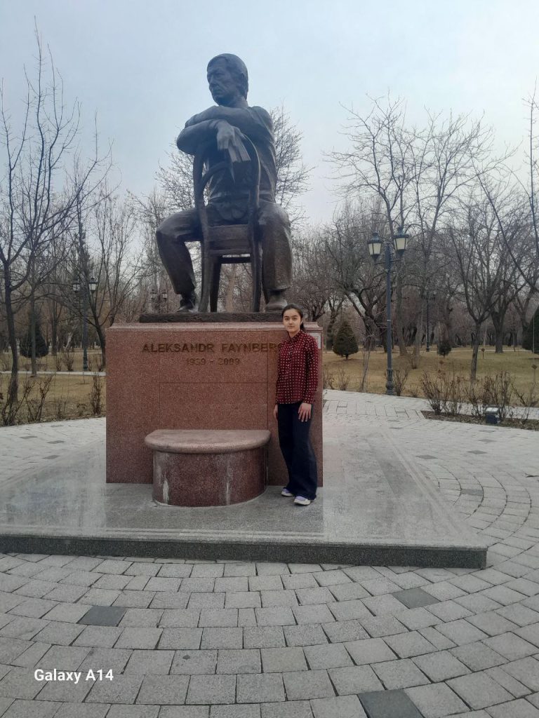 Young Central Asian woman in a red and black buttoned jacket standing in front of a giant statue of Alexandr Faynberg seated backwards in a chair. It's on a pedestal in a park with barren trees and lamps and grass. 