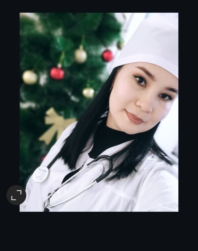 Young Central Asian woman in a doctor's white coat with a stethoscope leaning to the right. She's got long straight dark hair and dark eyes. 