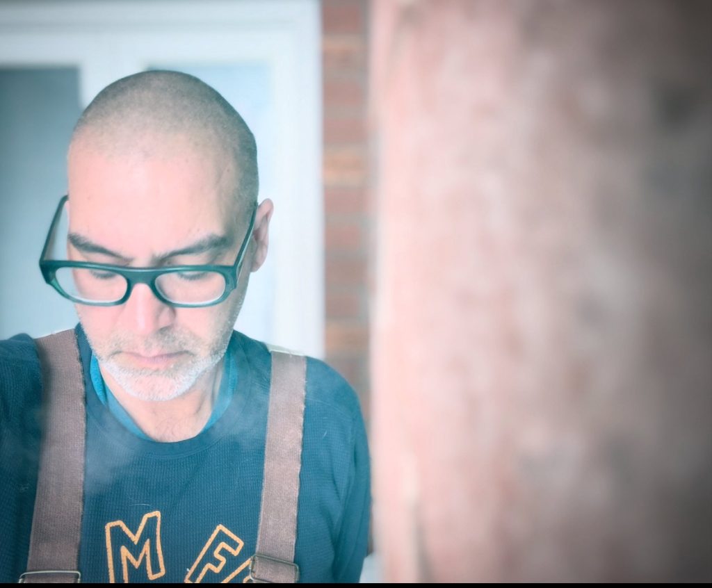 Older white man looking down at the floor. He's got reading glasses and brown suspenders and a blue tee shirt with some lettering. 