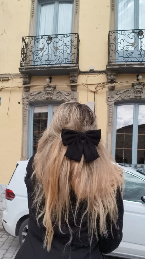 Blonde woman from behind with a black hair bow standing in front of a building with a yellow wall and decorative windows with light blue windowsills and brown filigree decor. 