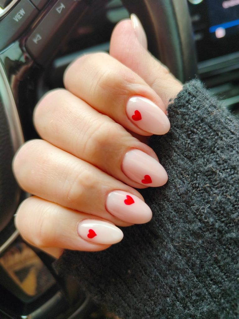 Closeup of a woman's hand with pink painted nails with little red hearts. She's got a gray sweater and is sitting inside a car. 