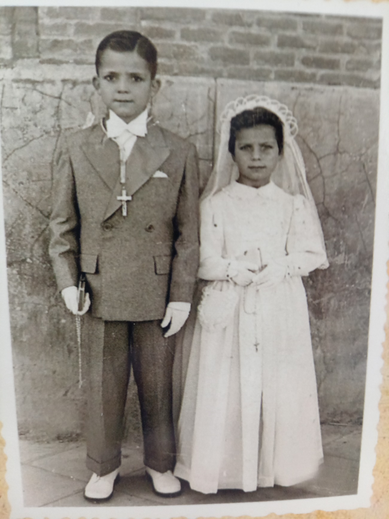 Black and white photo of a young boy in a suit with a cross and a young girl in a white lacey dress with a headdress in front of a brick wall. 