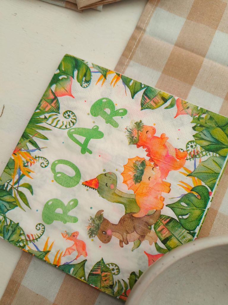 Kids' napkin with three cute green and orange dinosaurs, some foliage, a flower and fern, and "ROAR" in green font on a checkered tablecloth. 