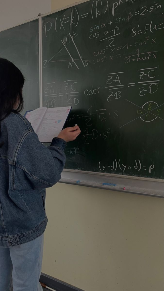 Teen Central Asian girl in a jean jacket with long dark hair writes mathematics on a green chalkboard. 