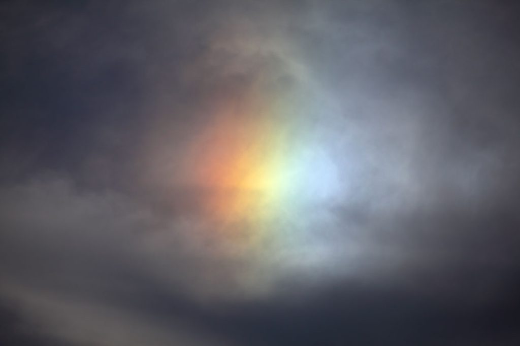 Rainbow clustered together, not an arc, visible in a gray cloudy sky. Called a "sundog." 