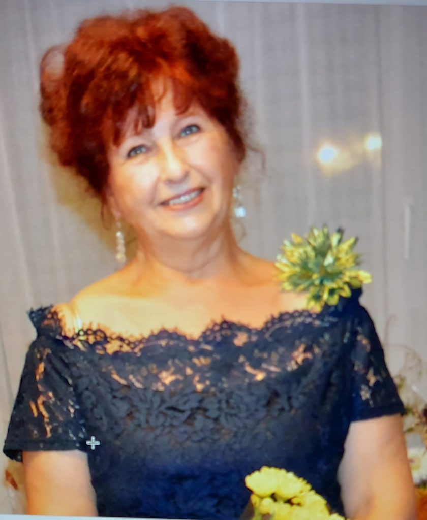 Light-skinned woman with red hair in a ruffled lacey black top with white dangling earrings and a yellow and green flower on her left shoulder.