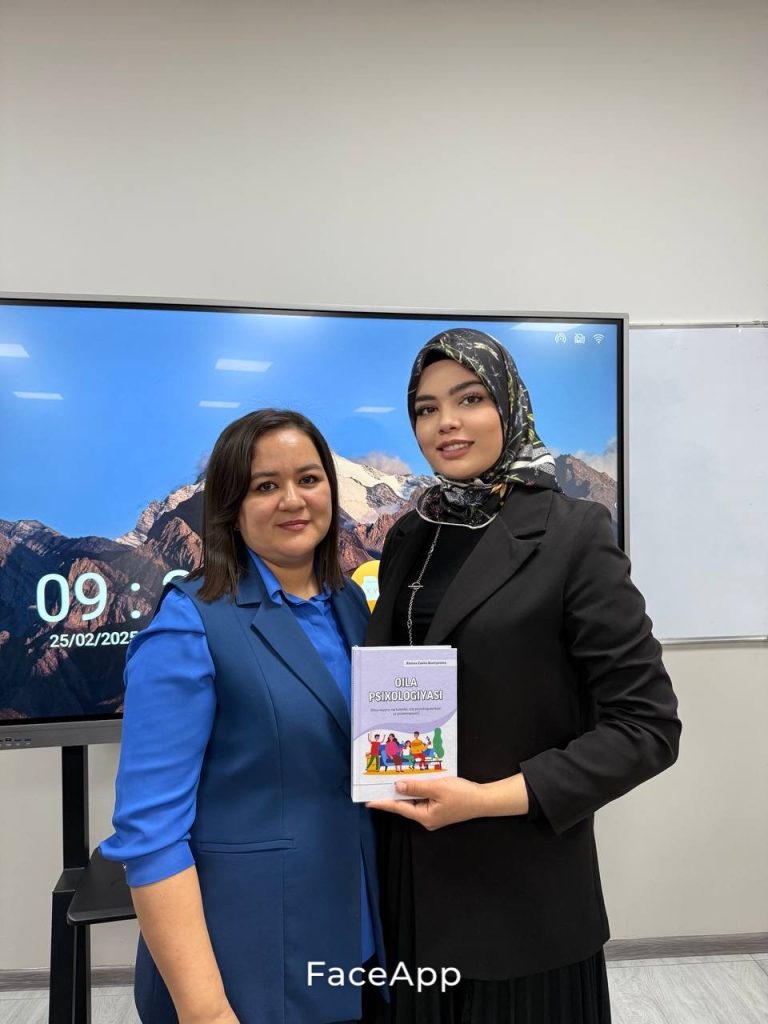 Two Central Asian women, one with a mostly black headscarf, black blouse, and black coat, and the other in a blue vest and top stand in a meeting room next to a computer screen.