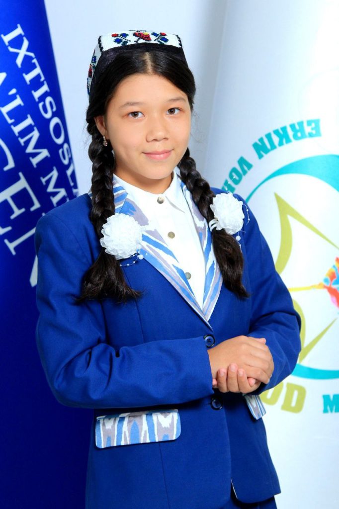 Young Central Asian woman with an embroidered headdress and braids of dark black hair and a blue jacket and white collared shirt (school uniform) standing in front of blue and white flags. 
