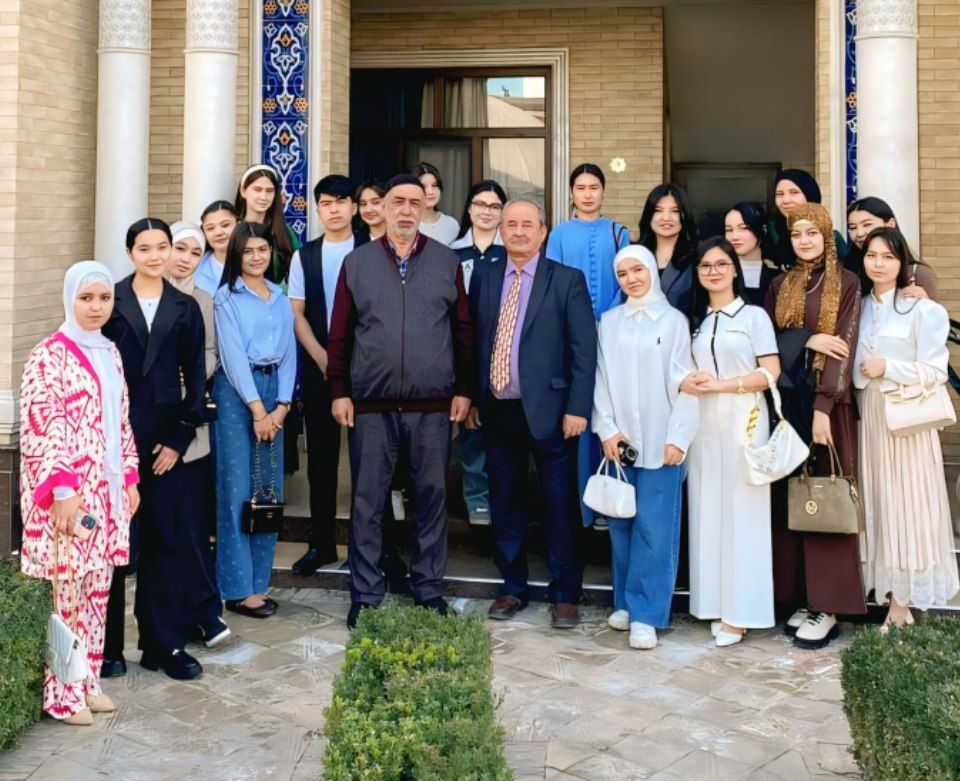 Large group of Central Asian college students and two older professors. Women in skirts and mostly dress shoes and blouses, some have headscarves. They're in front of a tall brick building that has columns and some Islamic style ornamentation.