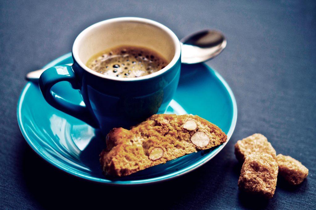 Blue, full cup of coffee on a light green saucer with some bits of biscotti.