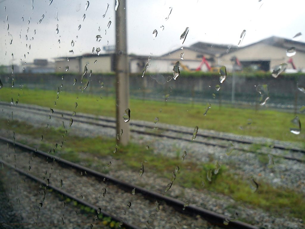 View out a train window of grass, train tracks, gray rocks, a pole, and distant houses on a rainy day