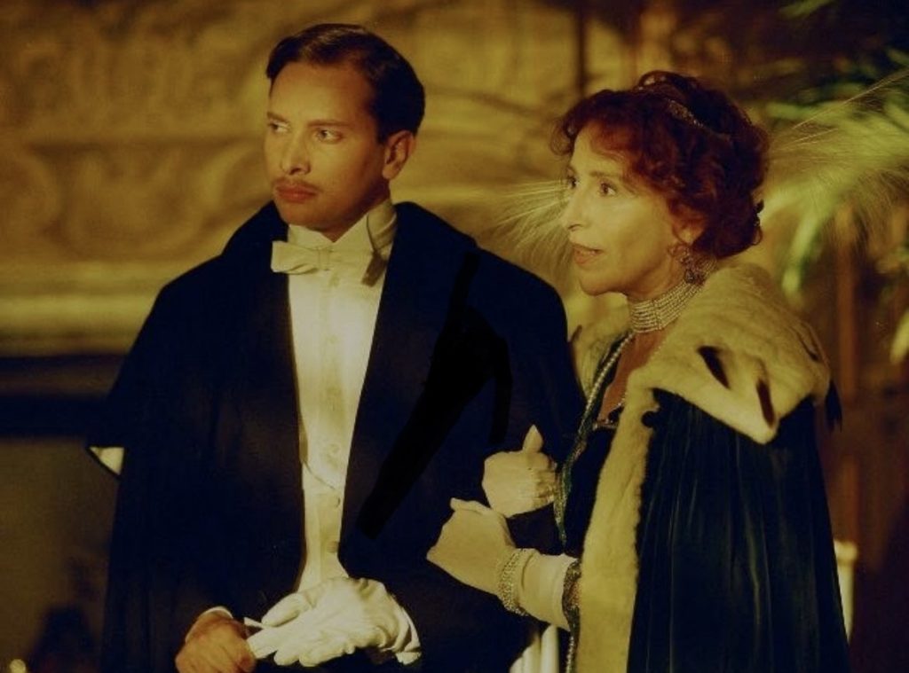 Young man and older woman dressed up in white gloves and suits and pearls in a movie.