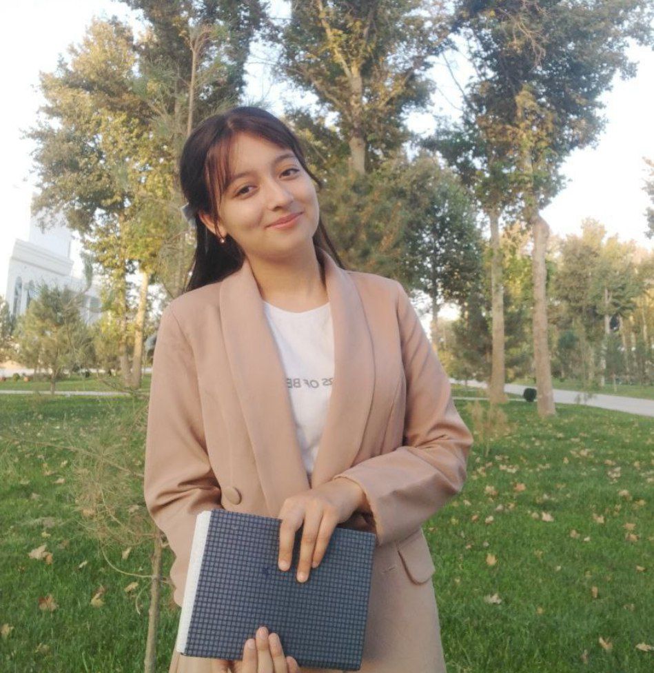 Young Central Asian woman holding a book and standing on a lawn near trees and large buildings. She's got a brown buttoned coat and white top.