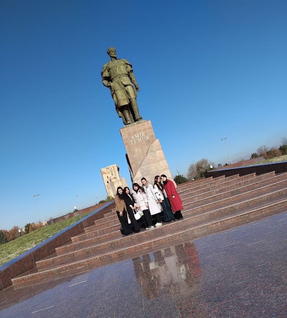 Group of Central Asian students in coats and black dress pants on the marble steps in front of a monument to Amir Temur. Sunny day, photo at an angle.
