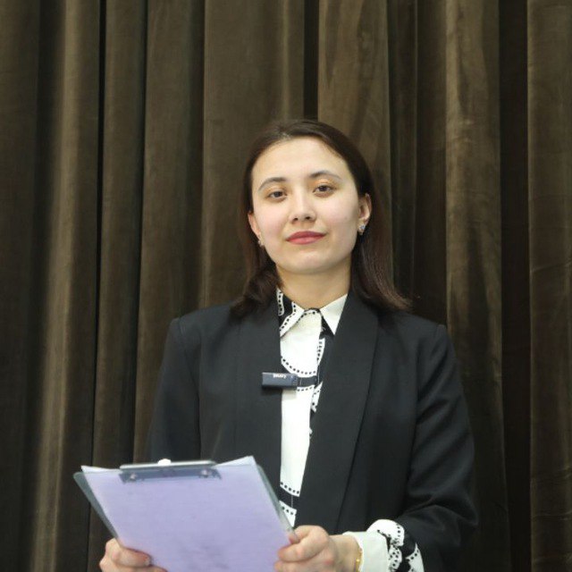 Central Asian young woman with dark hair and brown eyes and a white blouse and black coat in front of a brown curtain.
