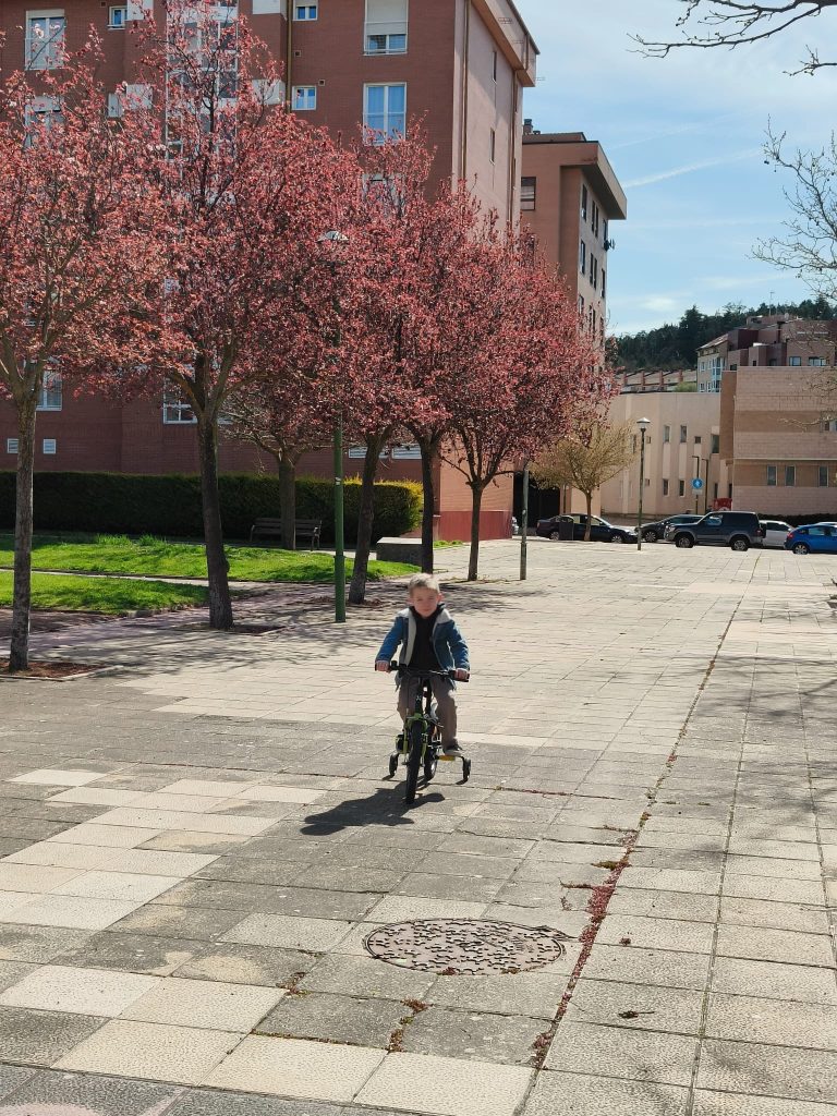 Boy riding a small bicycle on a path near blooming pink trees and lawns and peach colored brick apartment buildings. 