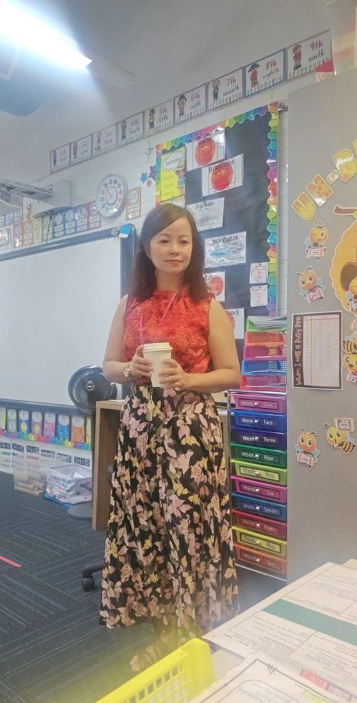 East Asian woman with straight dark hair, a red and orange blouse, black skirt with a leafy pattern, standing in an elementary school classroom.