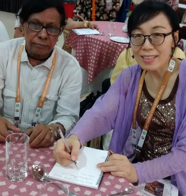 A South Asian middle aged man seated at a small table at a conference next to an East Asian woman. He has a white collared shirt and a lanyard and she has a purple coat over a gold and black blouse. She's taking notes, writing as he talks.