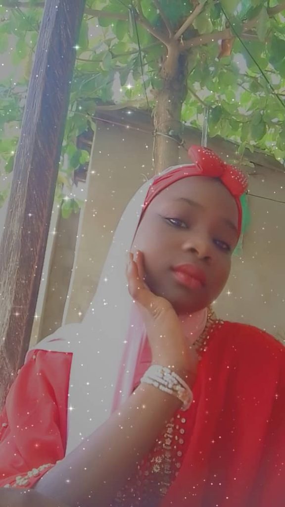 Young Black woman in a red blouse with a gold colored necklace, white veil, and a red headband and bow, in front of a brown wall or canvas and under a leafy green tree. 