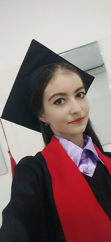 Young Central Asian woman in a graduation cap and gown with earrings, and a red sash and light purple silk collared shirt. 