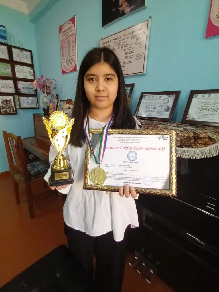 Young Central Asian woman with long straight dark hair stands in a room with a piano and a bunch of diplomas and awards on the wall. She's holding a certificate and a trophy.