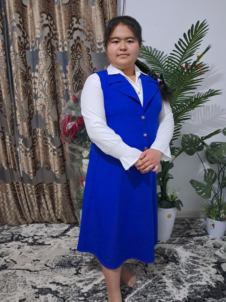 Central Asian woman in a blue vest and white collared blouse standing in front of a houseplant and a patterned curtain.