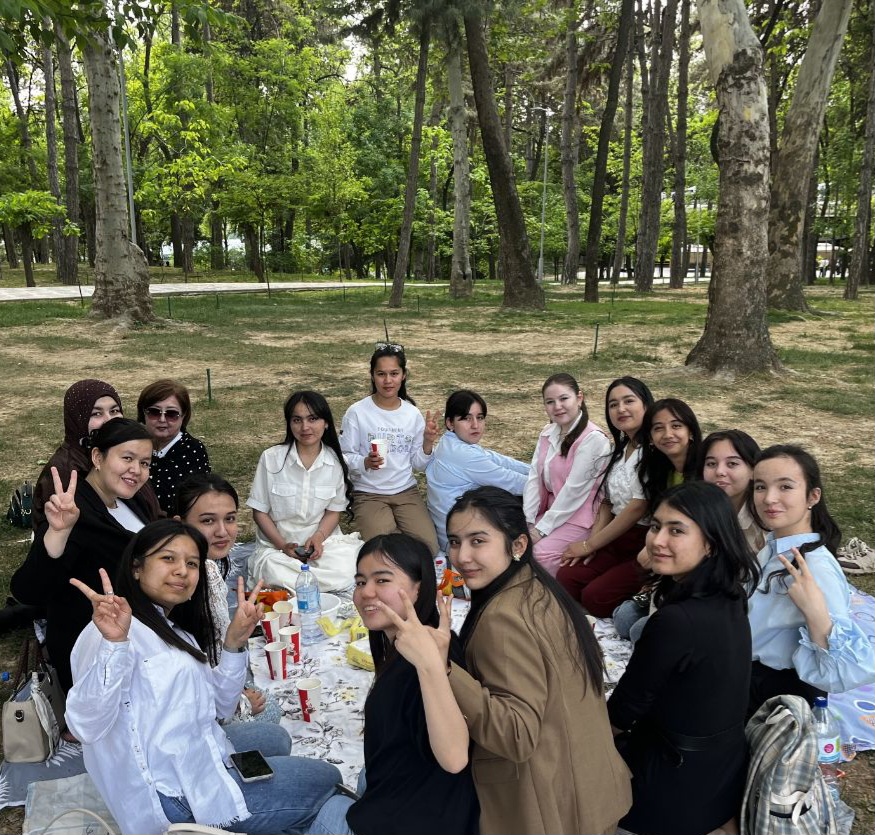 Variety of Central Asia students, young women, in dress clothes or uniforms, having an outdoor picnic near leafy trees.