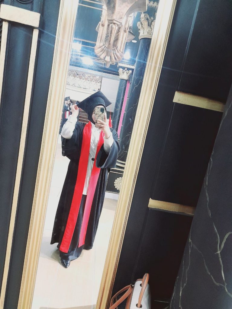 Young Central Asian woman in a black graduation gown and hat and red sash in a doorway in a room with black walls and a white framed mirror.