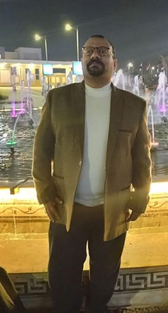 Middle aged Middle Eastern man in a brown coat standing in front of water fountains in a public square. 